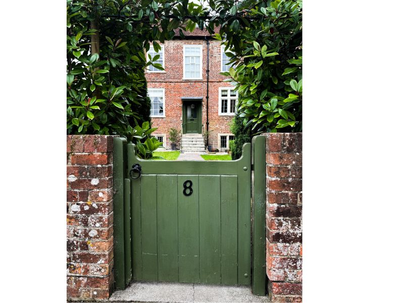 small green garden gate hung between two brick pillars and offering a view along a straight path to a house