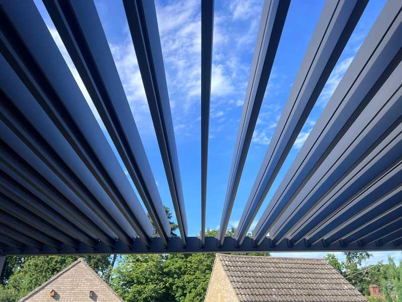 looking upwards towards the louvred roof of a modern garden pergola, louvres are partially open allowing a clear view of blue sky and white clouds